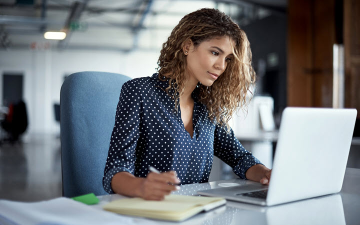 A woman seated at a desk in an office working on a laptop and writing in a notebook.