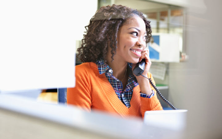 A smiling woman, in business attire, talking on a phone while sitting at a desk.